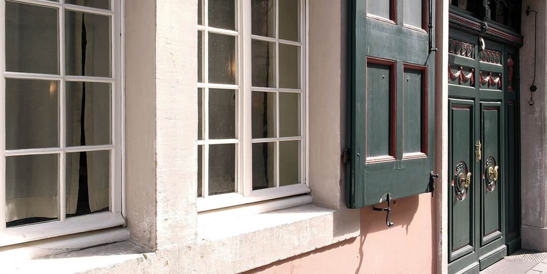 Nahaufnahme einer Hauswand mit zwei Fenstern in weißen Rahmen und einer grünen Doppelflügeltür